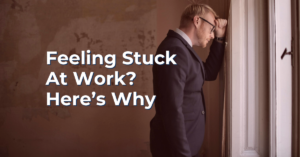 An image of a person in a corporate suit looking tired at their work with a caption that says, "Feeling Stuck At Work? Here's Why" for an article on why people feel stuck at work due to the zone of average and what they can do about it.