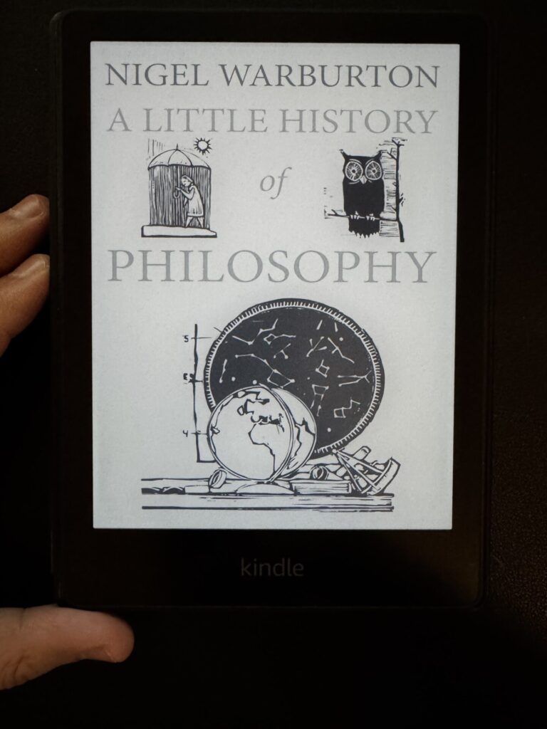 A book cover of Nigel Warburton's A Little History of Philosophy on Kindle and the hand of the author writing this book summary article.